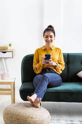 Junge Frau lächelt, während sie auf dem Sofa zu Hause sitzt und ein Mobiltelefon benutzt - GIOF11023