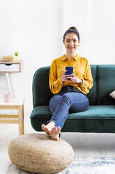 Lächelnde Frau, die ein Mobiltelefon benutzt, während sie zu Hause mit gekreuzten Beinen am Fußgelenk sitzt - GIOF11024