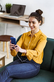 Junge Frau, die Musik hört, während sie ein Selfie mit ihrem Handy macht und zu Hause sitzt - GIOF11026