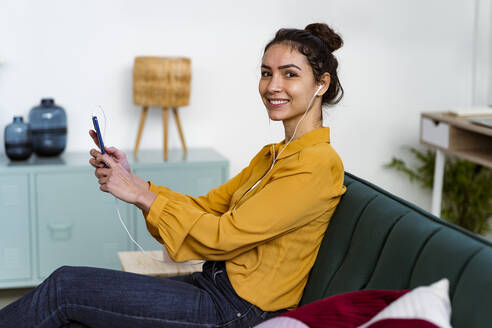 Lächelnde Frau mit In-Ear-Kopfhörern, die ein Mobiltelefon benutzt, während sie zu Hause auf dem Sofa sitzt - GIOF11027