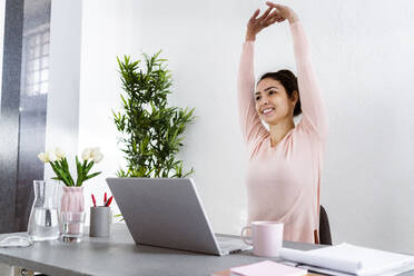 Junge Frau mit erhobener Hand, die einen Laptop benutzt, während sie im Heimbüro sitzt - GIOF11029