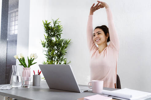 Young woman with hand raised using laptop while sitting at home office - GIOF11029