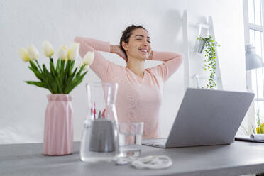 Young woman with hands behind head smiling while sitting at home office - GIOF11030