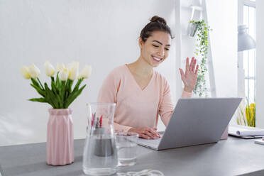 Young woman waving hand to video call on laptop while sitting at home office - GIOF11031