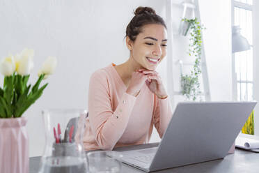 Young woman with hand on chin using laptop while talking with friend on laptop at home - GIOF11033