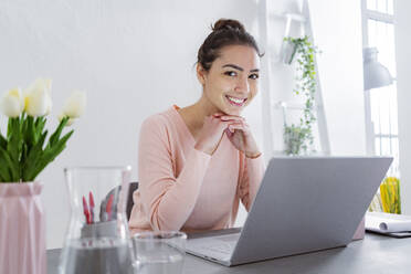 Young woman with hand on chin smiling while using laptop sitting at home office - GIOF11035