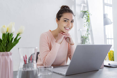 Junge Frau mit Hand auf dem Kinn lächelnd bei der Verwendung von Laptop sitzen im Büro zu Hause - GIOF11035
