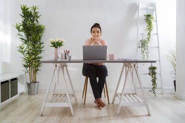 Lächelnde Frau mit Hand am Kinn, die einen Laptop benutzt, während sie zu Hause im Büro sitzt - GIOF11037