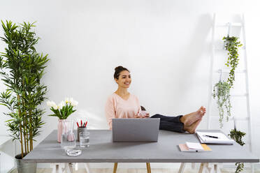 Smiling woman drinking coffee while sitting at home office - GIOF11038