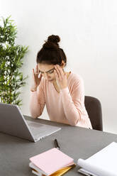 Young woman with head in hands sitting by laptop on table at home office - GIOF11043