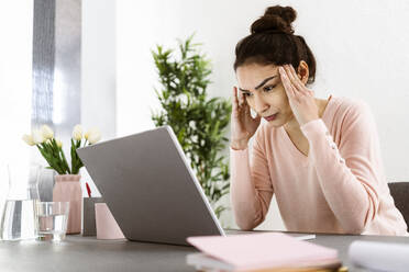 Woman looking at laptop while sitting with head in hands at home office - GIOF11044