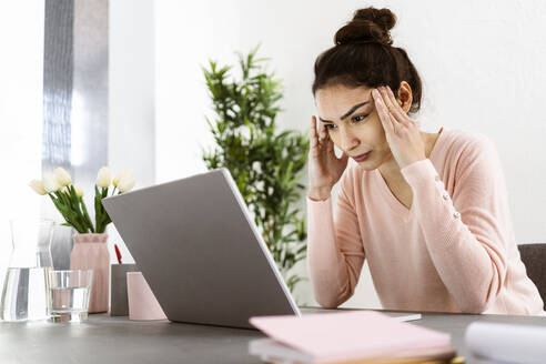 Frau, die auf einen Laptop schaut, während sie mit dem Kopf in den Händen im Heimbüro sitzt - GIOF11044