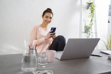 Lächelnde Frau, die ein Mobiltelefon benutzt, während sie zu Hause im Büro sitzt - GIOF11045