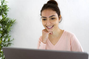 Smiling woman with hand on chin working on laptop while sitting at home - GIOF11046