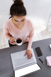 Frau mit Kaffeetasse bei der Arbeit am Laptop, während sie zu Hause sitzt - GIOF11049