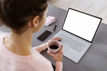 Junge Frau hält eine Kaffeetasse, während sie zu Hause am Laptop arbeitet - GIOF11050