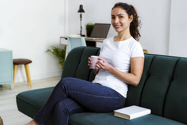Young woman smiling while drinking coffee sitting on sofa at home - GIOF11061