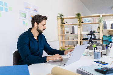 Männlicher Unternehmer, der ein Dokument betrachtet, während er einen Laptop im Büro benutzt - GIOF11070