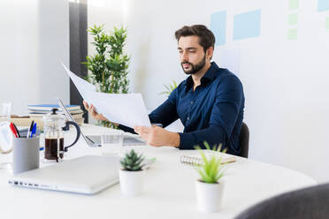 Männlicher Unternehmer, der Dokumente betrachtet, während er im Büro an der Wand sitzt - GIOF11072