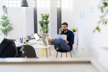 Young businessman discussing on smart phone while sitting with feet up at desk in office - GIOF11074