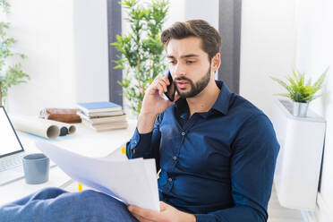 Businessman looking at documents while talking on smart phone in office - GIOF11075