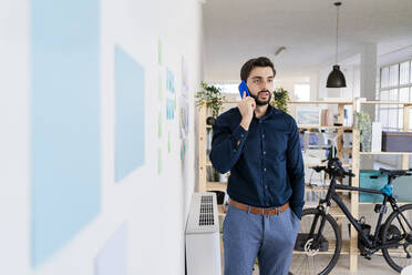 Männlicher Unternehmer, der mit der Hand in der Tasche am Smartphone spricht, während er im Büro an der Wand steht - GIOF11082