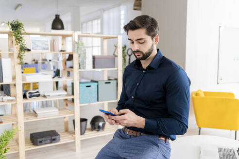Männlicher Unternehmer, der ein Smartphone benutzt, während er am Schreibtisch im Büro sitzt - GIOF11084