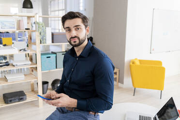 Junger Geschäftsmann sitzt am Schreibtisch und benutzt sein Smartphone im Büro - GIOF11085