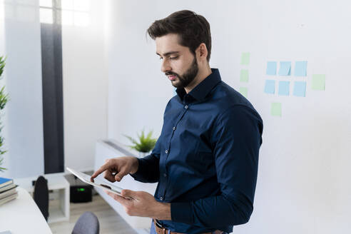 Männlicher Unternehmer mit digitalem Tablet an der Wand im Büro - GIOF11087