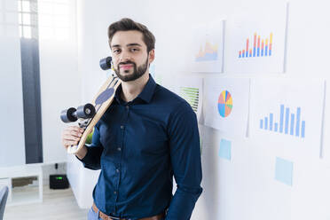 Männlicher Unternehmer mit Skateboard, während er im Büro an der Wand steht - GIOF11091