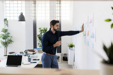 Junger Geschäftsmann, der Strategien analysiert, während er im Büro an der Wand steht - GIOF11096