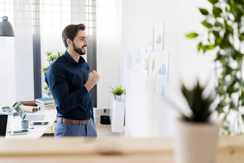 Fröhlicher männlicher Unternehmer ballt die Faust und betrachtet Strategien an der Wand im Büro - GIOF11100