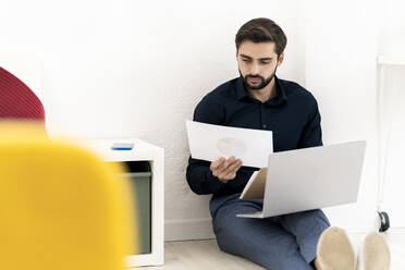 Junger Geschäftsmann analysiert Diagramme mit Laptop, während er im Büro an der Wand sitzt - GIOF11103