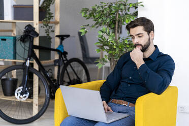 Nachdenklicher männlicher Unternehmer, der an einem Laptop arbeitet und auf einem gelben Stuhl im Büro sitzt - GIOF11105