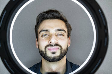 Handsome man seen through illuminated ring light against gray wall - GIOF11109