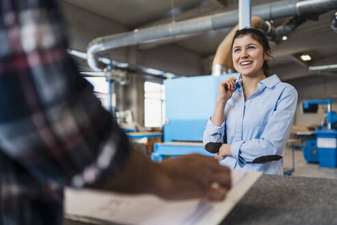 Lächelnde Geschäftsfrau bei der Arbeit mit einem Kollegen, während sie in der Industrie steht - DIGF14387