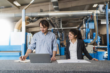 Businessman with laptop writing in book while standing by colleague at industry - DIGF14412