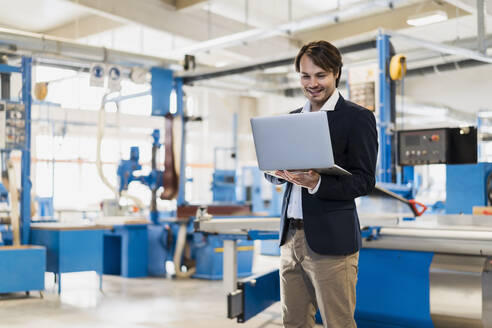 Junger Unternehmer, der einen Laptop benutzt, während er in der Industrie steht - DIGF14498