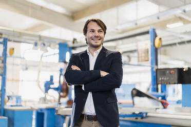 Smiling businessman with arms crossed looking away while standing at industry - DIGF14499