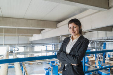 Lächelnde Geschäftsfrau mit verschränkten Armen an einem Geländer in der Industrie stehend - DIGF14504