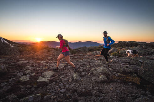 Mann und Frau beim Trailrunning mit Hund in den Bergen bei Sonnenaufgang - CAVF92318