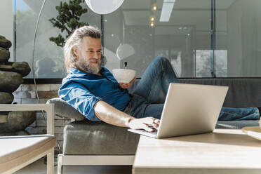 Mature man with a beard smiling and working with his laptop on a sofa - CAVF92485