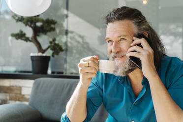 Mature man drinking a coffee and talking on the phone sitting on a sofa in his living room. Business concept - CAVF92489