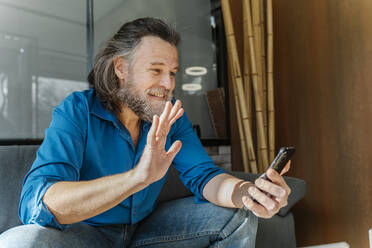 Älterer Mann mit Bart sitzt auf einem Sofa und schaut auf sein Smartphone - CAVF92490