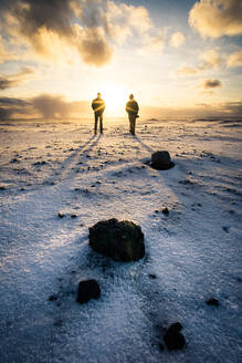 Zwei Menschen spazieren bei Sonnenaufgang am schwarzen Sandstrand in Island im Winter - CAVF92508