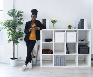 Afro female professional holding smart phone while leaning on rack at office - GIOF11113