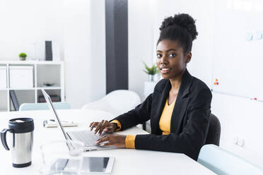 Junge Afro-Geschäftsfrau, die einen Laptop benutzt, während sie am Schreibtisch im Büro sitzt - GIOF11117