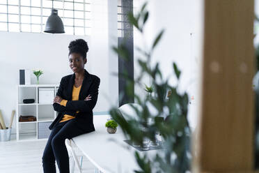 Smiling young Afro female entrepreneur with arms crossed sitting on desk in office - GIOF11136