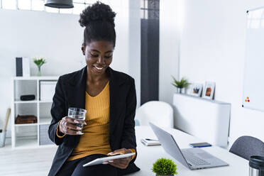 Lächelnde junge Afro-Fachfrau, die ein Glas Wasser hält, während sie ein digitales Tablet im Büro benutzt - GIOF11137