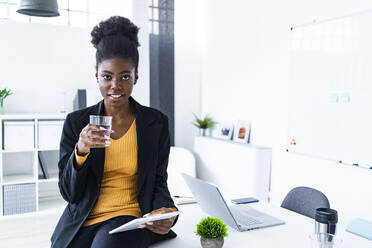 Young female Afro entrepreneur holding glass of water and digital tablet in office - GIOF11138